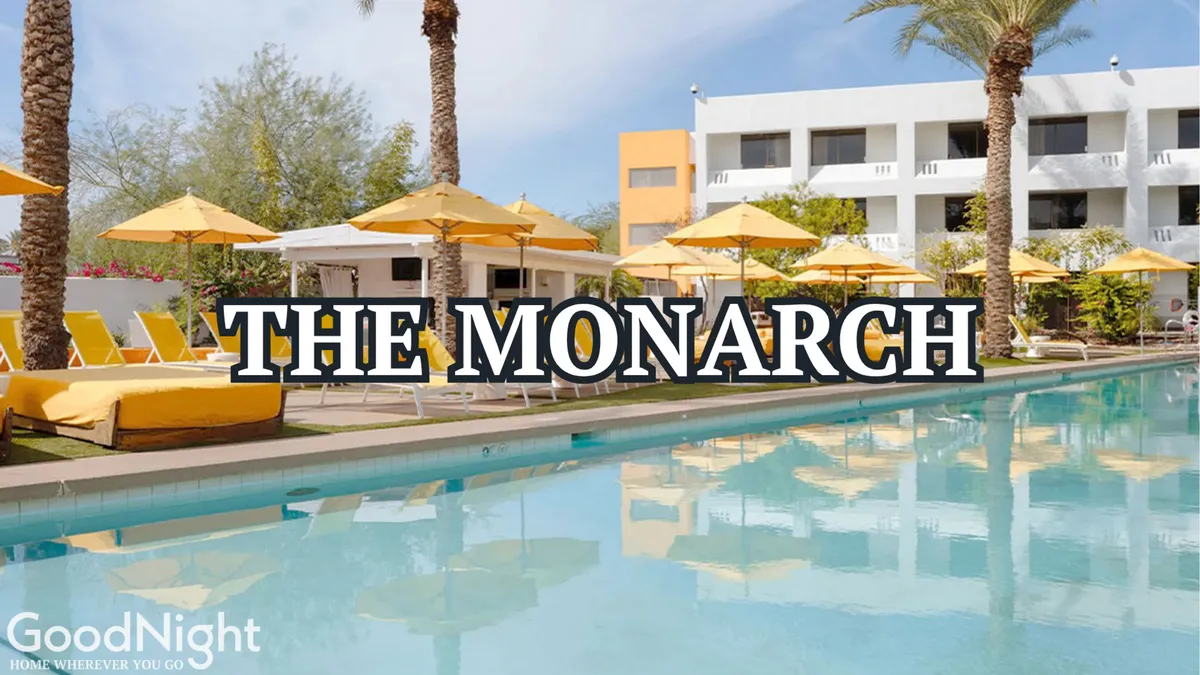 A luxurious resort-style swimming pool at The Monarch, complete with yellow umbrellas, lounge chairs, palm trees, and a hotel building in the background. The setting is bright and inviting, evoking a leisurely, vacation vibe.