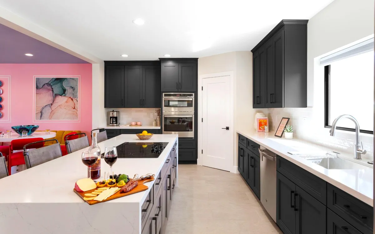 A cheerful, open kitchen and dining area with a central island topped with a cheese platter, sleek black cabinetry, and a brightly colored dining space beyond. The scene suggests shared meals, party prep, and easy-flow entertaining for groups.