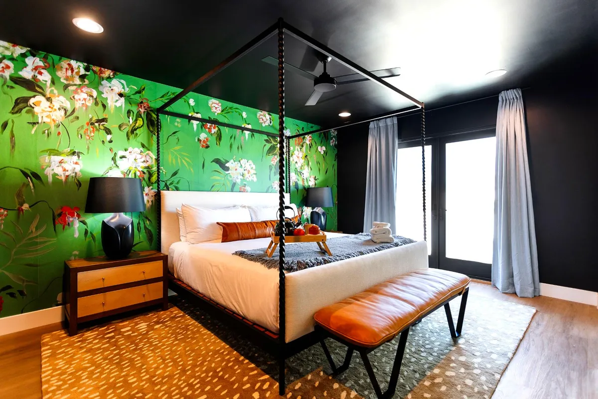 A chic bedroom featuring a four-poster canopy bed, vibrant floral wallpaper, and matching lamps on nightstands. A bench at the foot of the bed adds a practical touch, while the bright, airy feel suggests a stylish suite with high-end amenities.