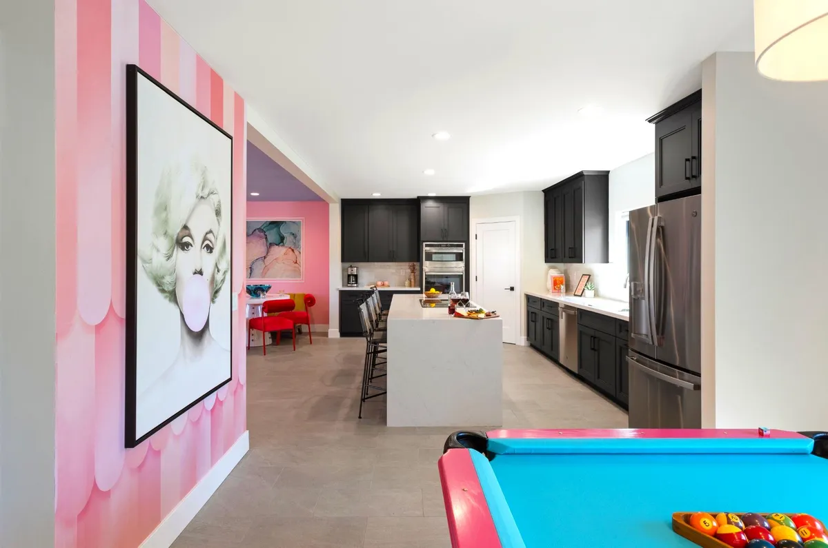 An interior shot of a playful game room featuring a modern pool table with colorful balls, a kitchen island, and a large artwork of a woman blowing bubble gum, infusing the space with vibrant, Marilyn Monroe-inspired flair.