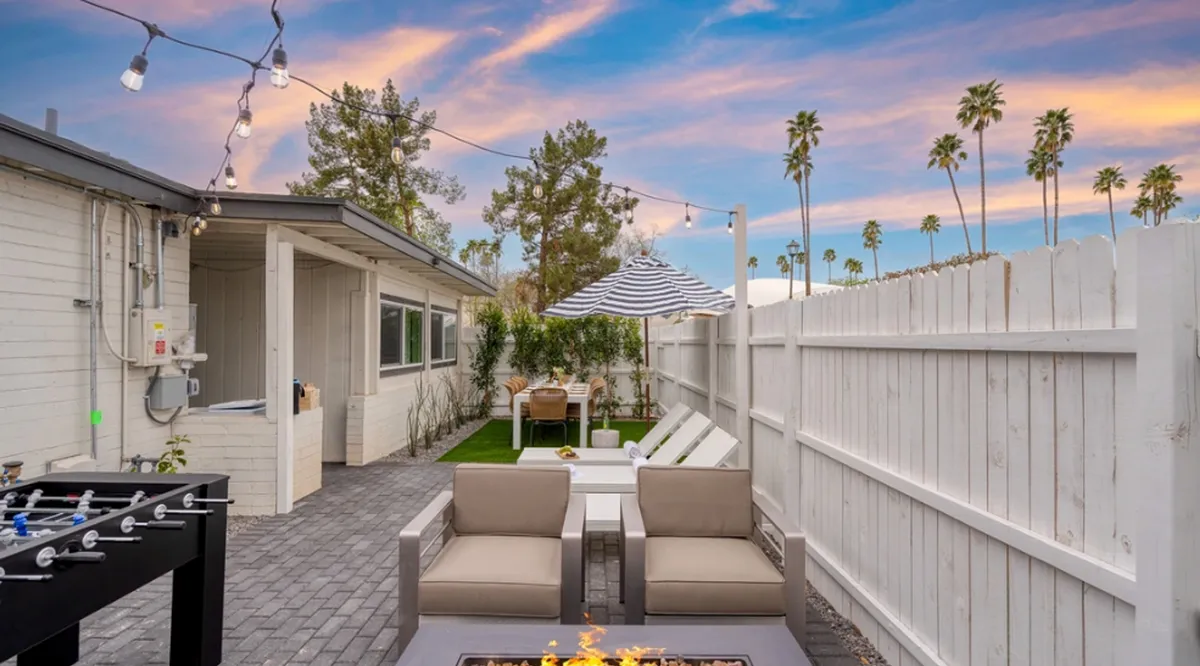 This image captures an inviting outdoor space featuring a fire pit surrounded by comfortable seating, a dining table under an umbrella, and string lights overhead. The ambiance is cozy and sociable, perfect for gatherings at dusk.