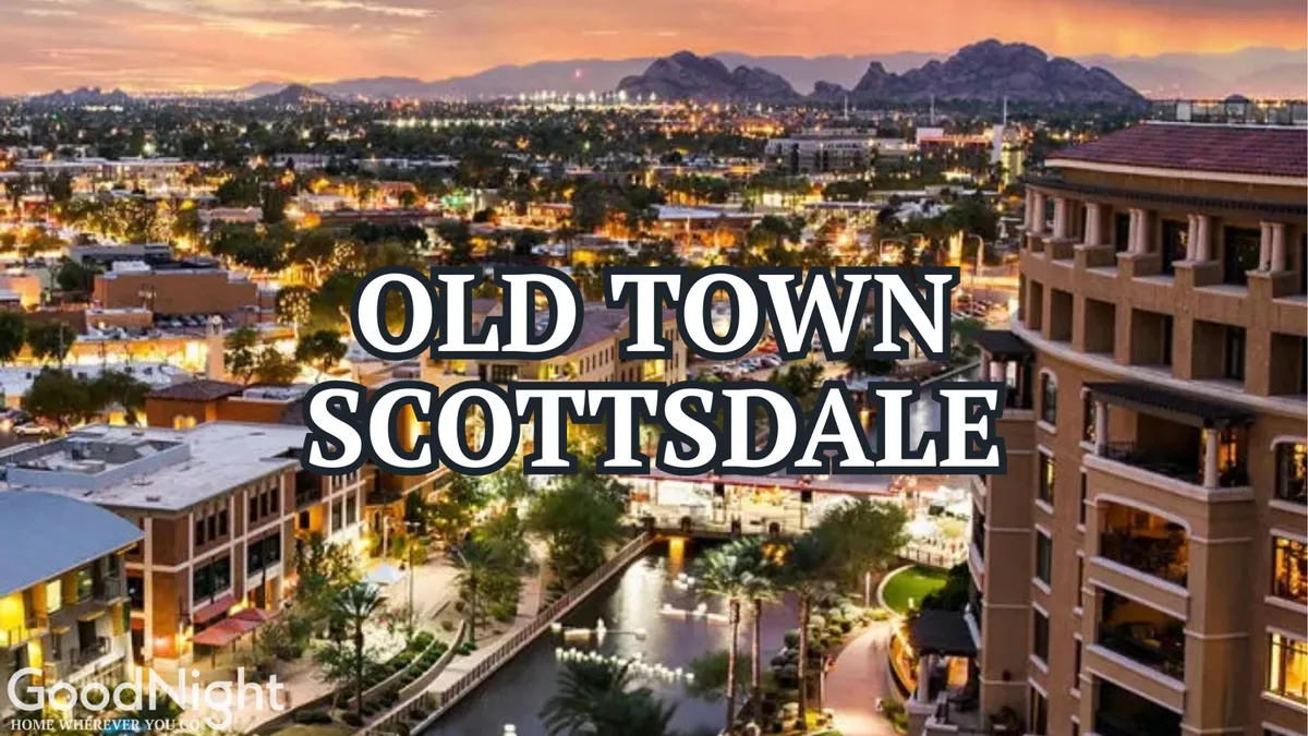A vibrant exterior view featuring the iconic Old Town Scottsdale sign illuminated at dusk, city lights sparkling in the background, with a scenic canal running alongside and mountains in the far distance. People can be seen strolling, shopping, and dining at outdoor cafes.