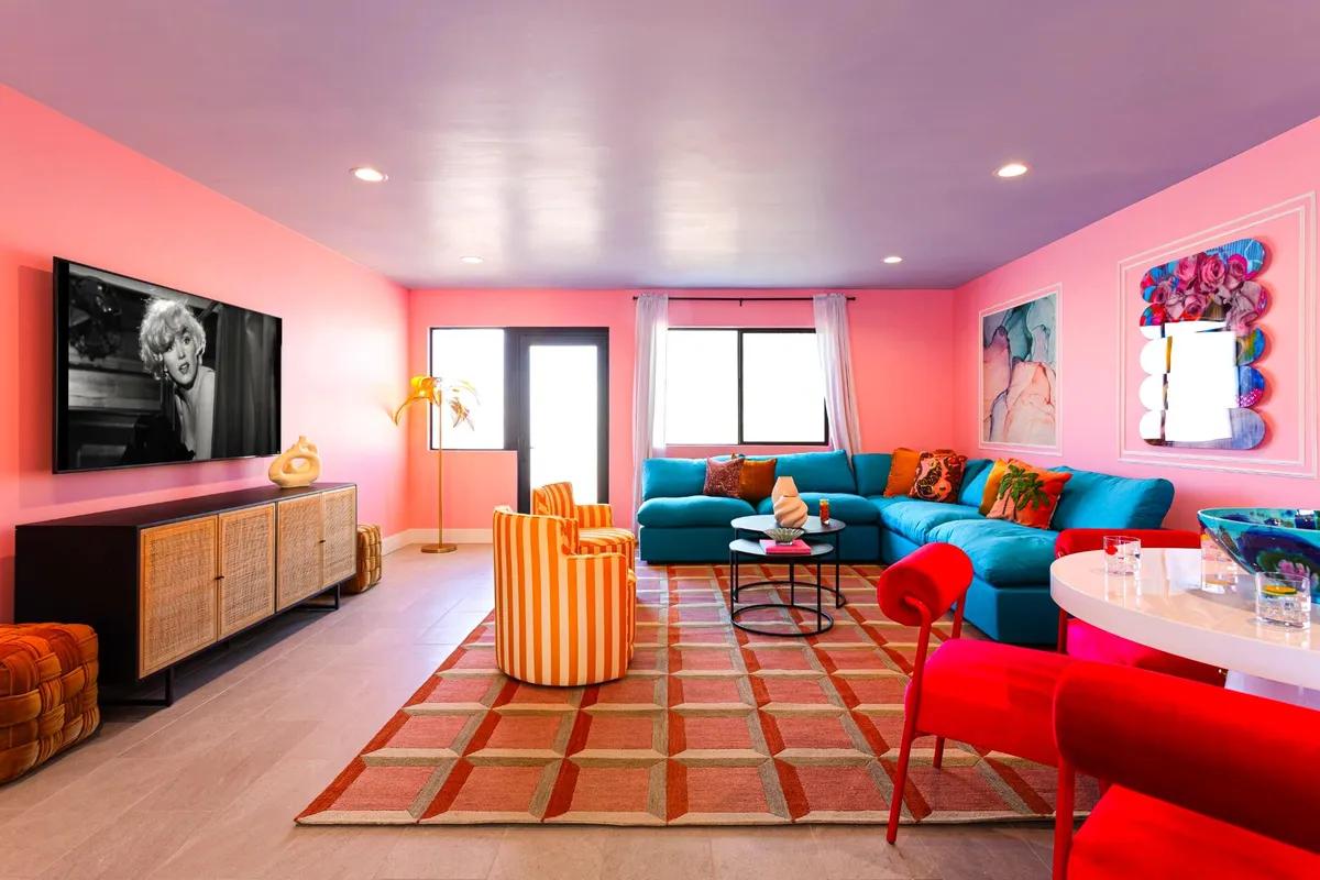 A bright, joyful living room featuring vivid pink walls, a teal sofa, striped accent chairs, a patterned rug, and a vintage television. The energetic decor is designed for lively gatherings, and the colorful setting makes an ideal backdrop for group photos and celebrations.