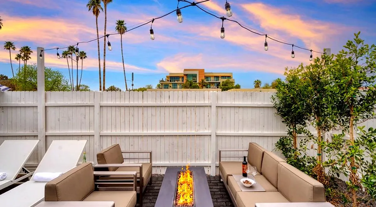 An inviting backyard scene featuring a fire pit surrounded by comfortable outdoor seating, illuminated by hanging lights and framed by palm trees, creating a cozy atmosphere for group gatherings or unwinding after a night out.