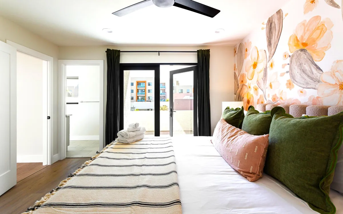 A calming bedroom with a large bed dressed in plush linens and decorative pillows, set against a floral accent wall. Glass doors open to an outdoor space—suggesting access to a private balcony for enjoying fresh air and morning sun.
