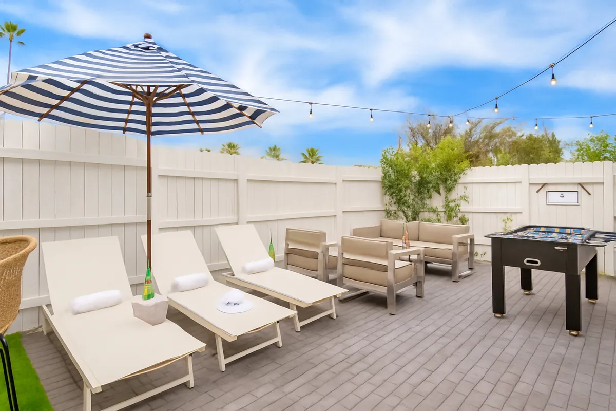 A cheerful backyard scene with striped umbrella, plush lounge chairs, a shaded seating area, and a foosball table. The space is designed for sunbathing, playful competition, and unwinding in style.